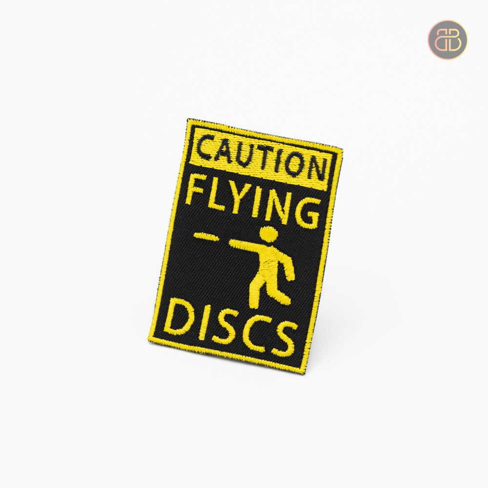 Discgolf patch with the text CAUTION FLYING DISCS and a person throwing a disc being showed on the patch