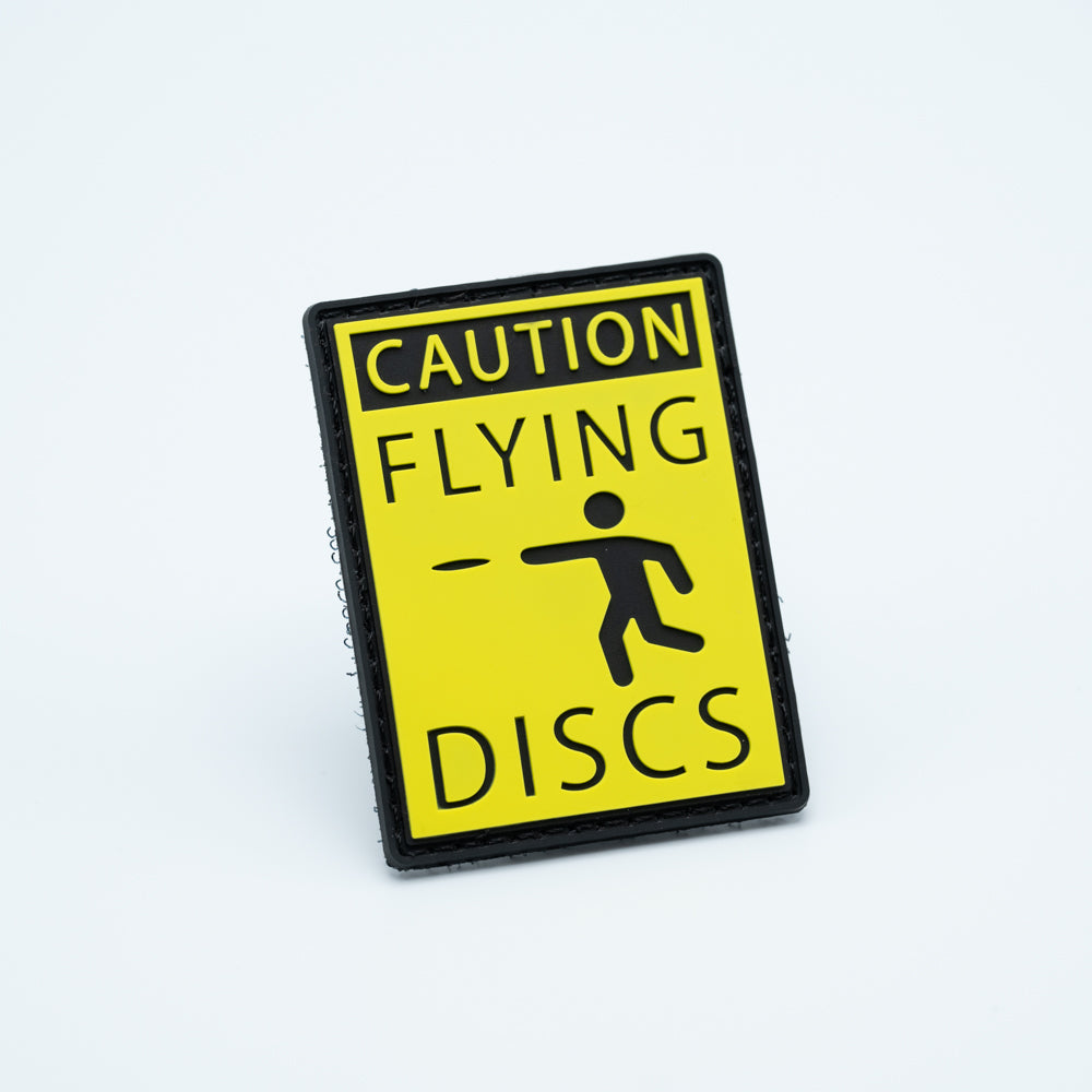 A PVC disc golf patch in yellow with the text CAUTION FLYING DISCS and a cartoon throwing a disc golf disc