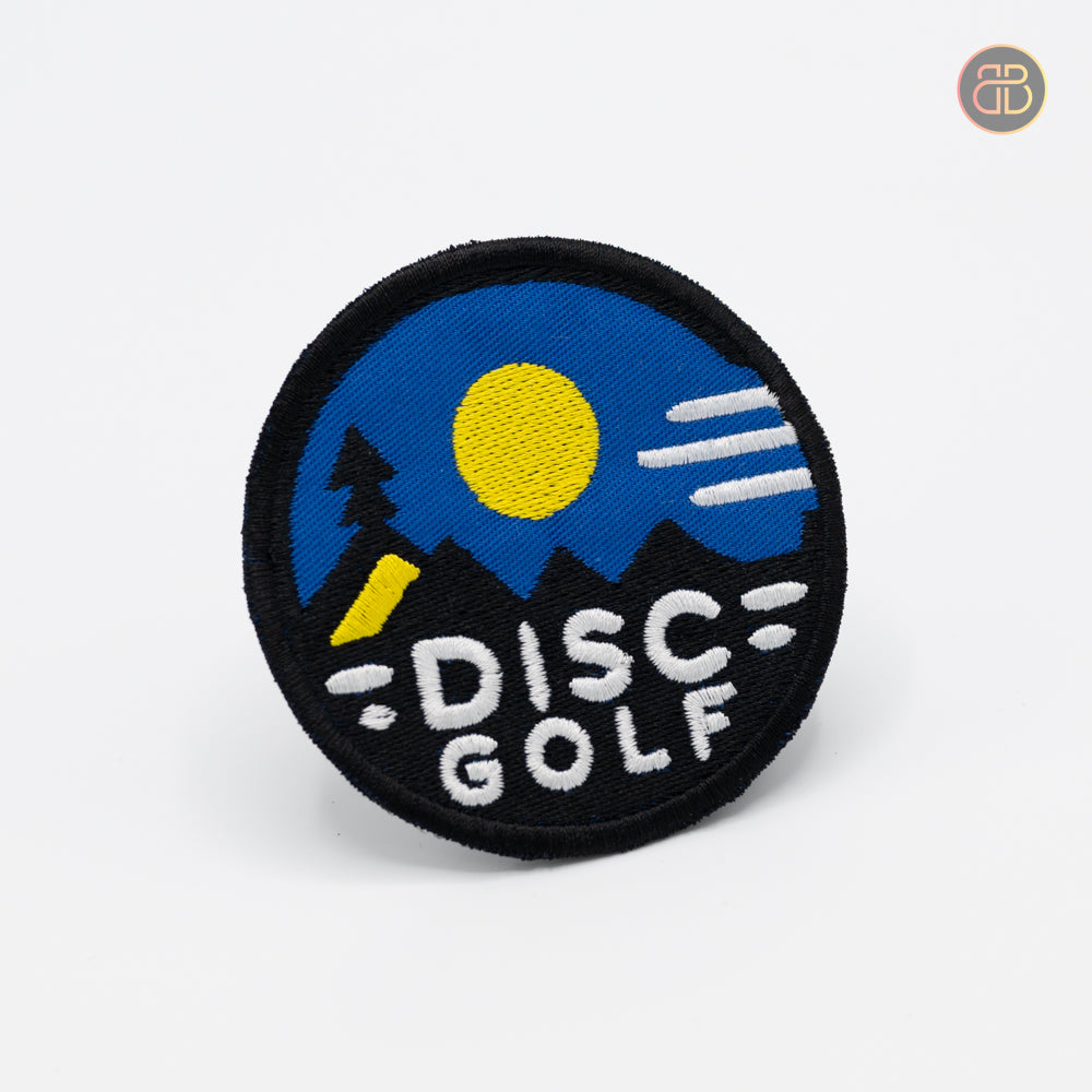 Discgolf patch with a sun and some mountains and a tree and the text DISC GOLF