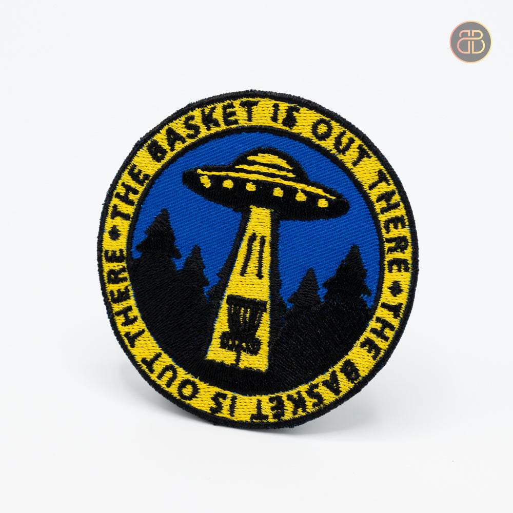 Discgolf patch with a ufo searching for a discgolf basket with the text "the basket is out there"