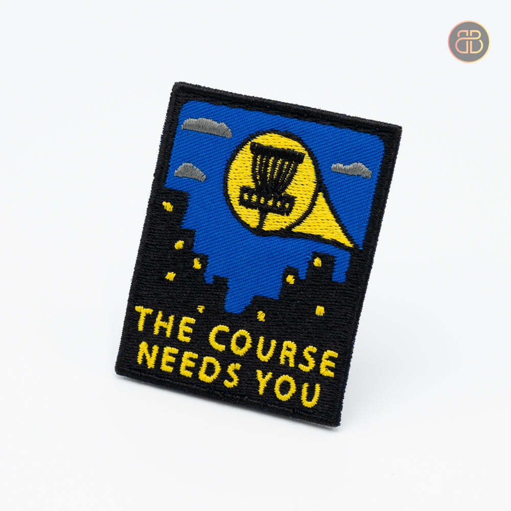 Discgolf patch. The text "The Course Needs you" in yellow. Also black houses with windows that are yellow. A disc golf basket is also shown in the picture.